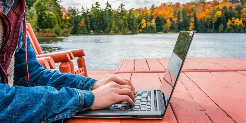 Jemand sitzt auf einer roten Sitzgarnitur an einem See und tippt auf einem Laptop