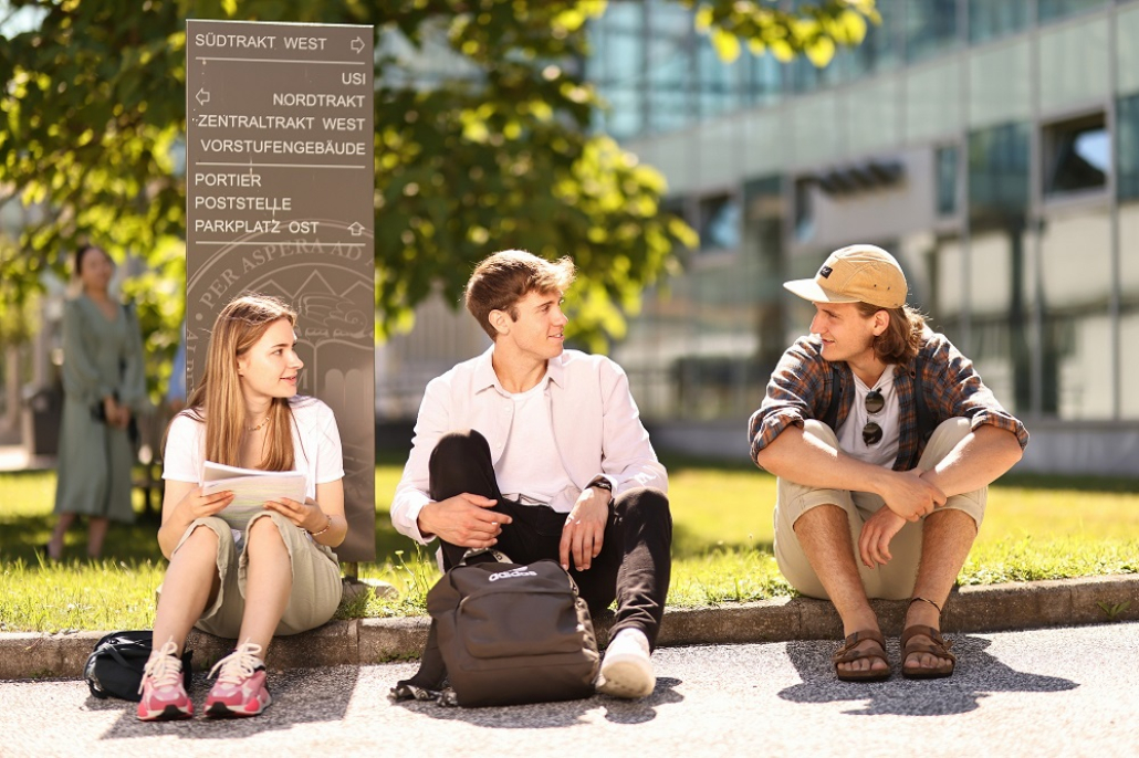 Studierende sitzen vor einem Orientierungsschild an einer Wiese auf dem Campus der Universität Klagenfurt.