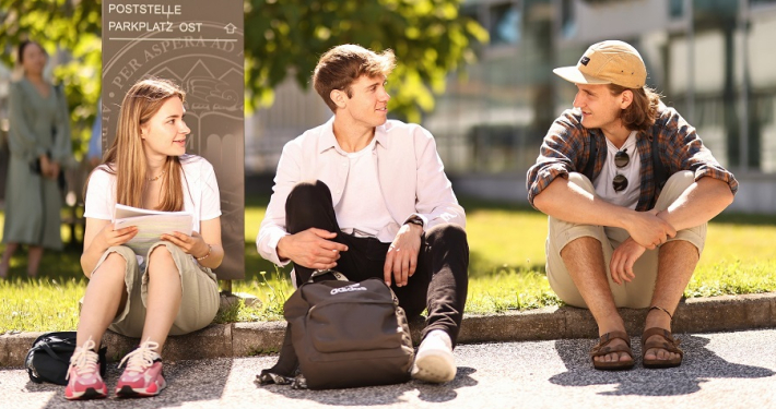 Studierende sitzen vor einem Orientierungsschild an einer Wiese auf dem Campus der Universität Klagenfurt.