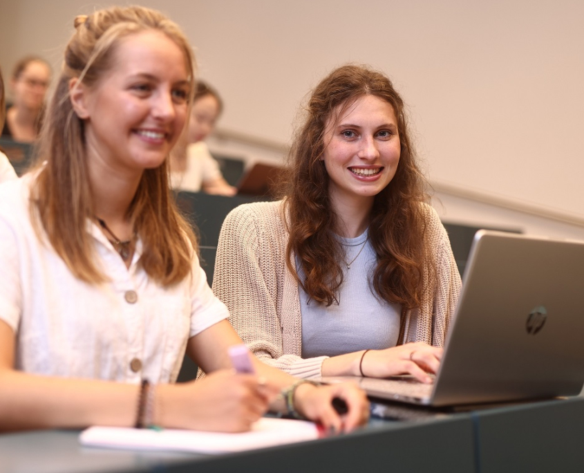 Zwei Studentinnen sitzen mit einem Laptop in einer Sitzreihe im Hörsaal.