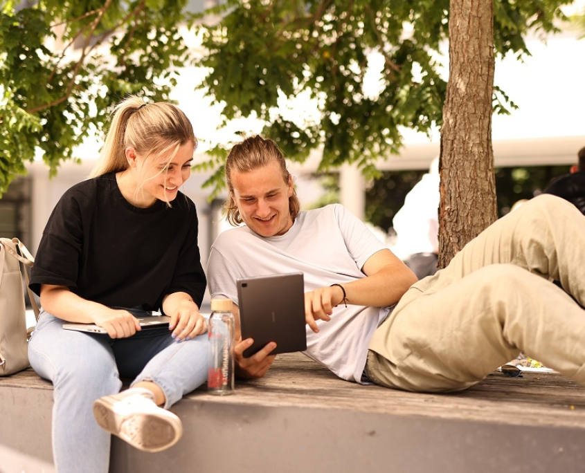 Die Sitzgelegenheiten im Außenbereich des Campus der Universität Klagenfurt werden im Sommer häufig zum gemeinsamen Lernen genutzt. | aau/Daniel Waschnig Zwei Studierende sitzen mit Touchpad und Laptop auf einer steinernen Sitzgelegenheit unter einem Baum.